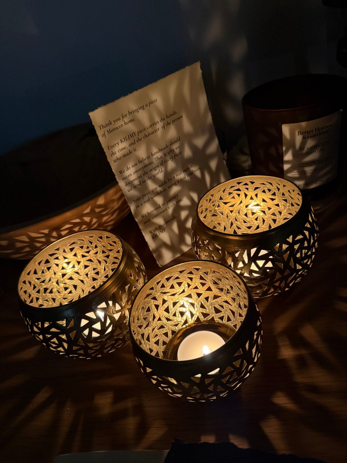 Three hand-engraved brass tealight holders lit on a wooden table at night, beside the Kilimy thank-you letter